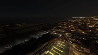 A coastal airport at night shows an illuminated parking lot, terminal buildings, parked light aircraft, and runway lights beside surrounding neighborhoods.