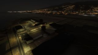A coastal airport at night shows illuminated buildings, parking spaces, and runway lights beneath a hillside dotted with city lights.
