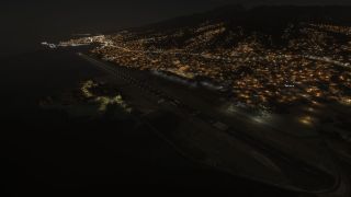 An illuminated coastal runway lies beside dark water at night, with dense city lights climbing the surrounding mountain slopes.