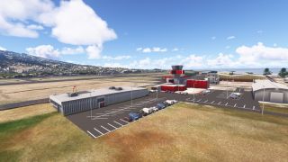 A red-and-gray control tower stands beside airport parking lots, with runways, hangars, hillside buildings, and the sea beyond.