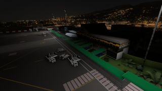 An airport terminal overlooks an apron at night, with green covered walkways, parked ground vehicles, and hillside lights in the background.