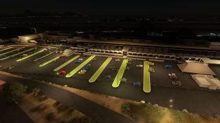 An airport terminal overlooks a lit parking lot at night, with rows of cars, narrow green islands, and covered walkways.