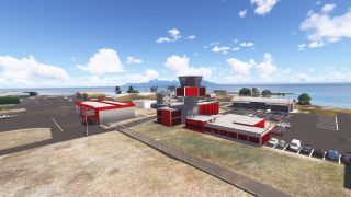 A red-and-gray control tower stands beside airport hangars and parked cars, with coastal waters and a mountainous island beyond.