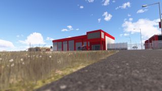 A red airport fire station stands beside a fenced access road, with a fire truck parked outside its gray garage doors.