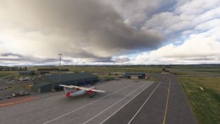 A red-and-white twin turboprop sits beside airport hangars, with apron markings, a control tower, and heavy clouds visible.