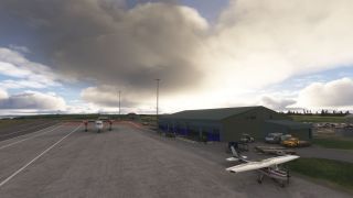 A high-wing light aircraft and twin turboprop sit on an airport apron beside a green building and service vehicles under large clouds.