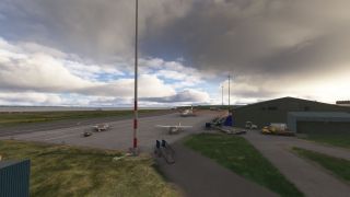 Light aircraft and a twin turboprop sit on an airport apron beside green hangars, service vehicles, and tall light poles under heavy clouds.