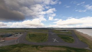 A coastal airport has a runway marked with white crosses, adjoining taxiways, hangars, and parked aircraft beneath scattered clouds.
