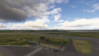 A small airport control tower stands beside parked cars and a red-and-white mast, with runways extending across grass beneath scattered clouds.