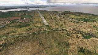 Runway 36 extends toward the shoreline, with adjacent taxiways, scattered buildings, and grassy fields beneath a cloudy sky.