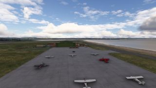Several light aircraft, including a red biplane, sit on a concrete apron beside green hangars and a shoreline taxiway.