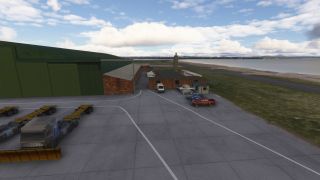 A green hangar and brick service buildings stand beside parked trucks, trailers, and cars near a shoreline under scattered clouds.