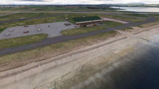 Several light aircraft sit on an apron beside a green-roofed hangar, with taxiways and runways bordering a sandy shoreline.