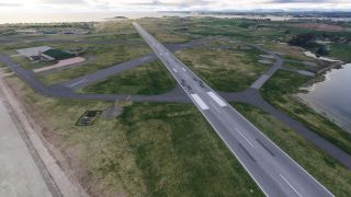 A coastal airport has a long paved runway, intersecting taxiways, green-roofed hangars, and grassy areas bordered by water.