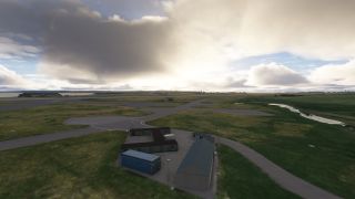 Small airport buildings and a blue shipping container stand beside paved taxiways, grassy fields, and a winding stream beneath heavy clouds.