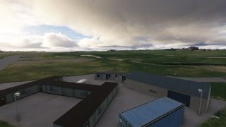 Low airport buildings, a hangar, and a blue shipping container stand beside grassy airfield pavement under thick clouds.