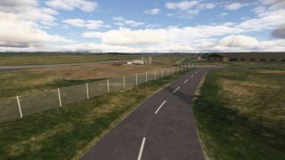 A paved road curves alongside an airport perimeter fence, with cylindrical storage tanks, grassy fields, and low buildings beneath scattered clouds.