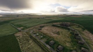 Small buildings line narrow roads amid green coastal fields, with an airfield and water stretching beneath low clouds in the distance.