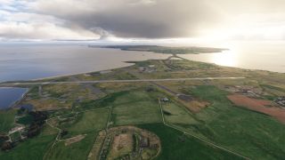 Intersecting runways and taxiways span a coastal airfield surrounded by green fields, scattered buildings, and water beneath low clouds.