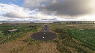 Runway 06 extends from a circular paved turnaround toward the water, with grassy surroundings, scattered hangars, and low clouds overhead.