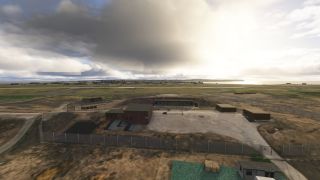 A fenced compound contains brick buildings and earth-covered shelters beside an airfield, with coastal water visible beneath heavy clouds.