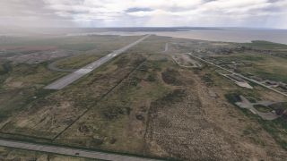 Runway 36 extends toward the coastline, with connecting taxiways, grassy fields, scattered buildings, and roads beneath a cloudy sky.