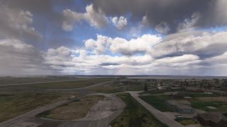A runway and scattered airport buildings sit beneath layered clouds, with a faint rainbow visible near the horizon.