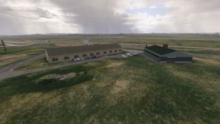 Two low airport buildings stand beside parked cars and grassy fields, with paved taxiways extending beneath a cloudy sky.