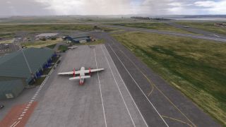 A red-and-white twin turboprop sits on an apron beside a green-roofed terminal, with runways and smaller aircraft beyond.