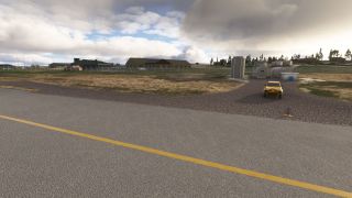 A yellow vehicle sits beside storage tanks near a taxiway with a yellow line and blue edge lights beneath cloudy skies.