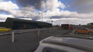 A low airport terminal faces a car park with orange and yellow cars, white bollards, and flagpoles beneath cloudy skies.
