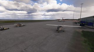 A high-wing light aircraft, a biplane, and a twin turboprop sit on the apron beside a terminal under cloudy skies.