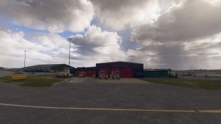 Two fire engines stand outside an airport fire station with red bay doors, beside service vehicles beneath heavy clouds.