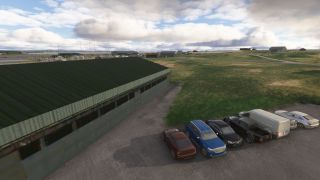 Cars, a pickup truck, and a white van sit beside a green-roofed hangar, with grassy airfield grounds stretching toward scattered buildings.