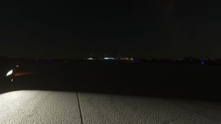 Bright lights illuminate the airport pavement at night, with blue taxiway lights and lit buildings visible in the distance.