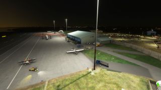 Floodlights illuminate an airport apron at night, where a biplane, light aircraft, and twin turboprop sit beside a terminal and car park.