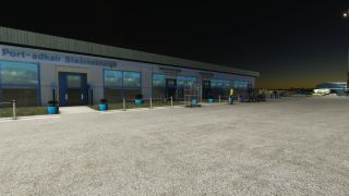 The airport terminal stands beside an illuminated apron at night, with blue-framed doors, potted shrubs, baggage carts, and ground personnel.