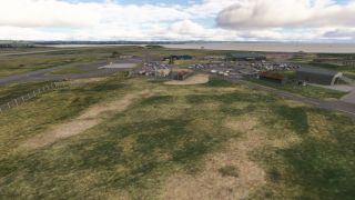 A coastal airfield has parked light aircraft, hangars, fenced fuel tanks, and car parks beside grassy taxiways under scattered clouds.