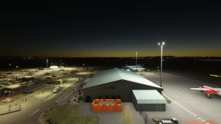 Floodlights illuminate an airport terminal, a car park, and a red-and-white aircraft on the apron beneath a dark sky.