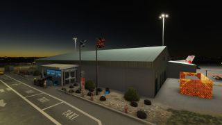 A floodlit airport terminal stands beside marked taxi bays, with Scottish and British flags outside and an aircraft tail visible behind.
