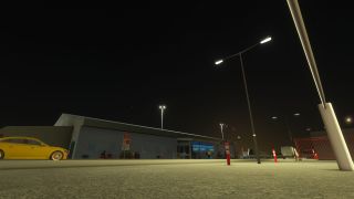 Streetlights illuminate a low airport building at night, with a yellow car, red-and-white bollards, and a red gate outside.