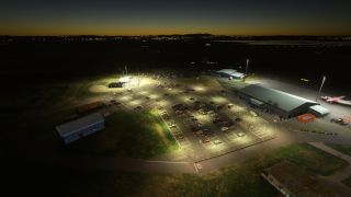 An airport terminal and car park sit under floodlights at twilight, with a red-and-white aircraft parked on the adjacent apron.