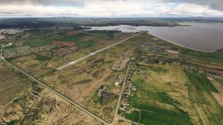 A coastal airport runway stretches toward the water, with connecting taxiways, nearby buildings, patchwork fields, and scattered houses beneath cloudy skies.