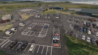 An airport parking lot contains cars and vans beside hangars, with light aircraft, taxiways, and a runway beyond.