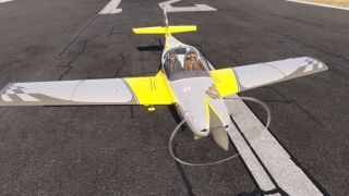 A yellow and gray low-wing aircraft sits on a cracked runway with two occupants, checkered wingtips, and a spinning propeller.