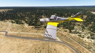 A yellow and gray light aircraft with checkered markings flies low above forest and dry grassland beside a winding paved road.