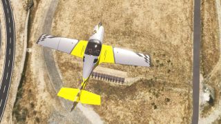 A yellow and silver aircraft with checkered wingtips flies above dry grassland and roads, viewed directly from overhead.