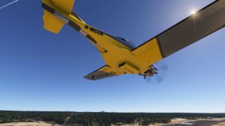 A yellow and black single-engine aircraft flies overhead, showing its underside and spinning propeller above a forest-bordered airfield.