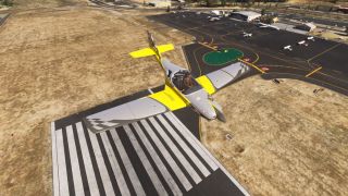 A yellow and gray single-engine aircraft flies above runway threshold markings, with parked aircraft and hangars across the apron behind it.