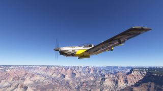 A yellow and silver single-engine aircraft with checkered markings flies above layered canyon cliffs beneath a clear blue sky.
